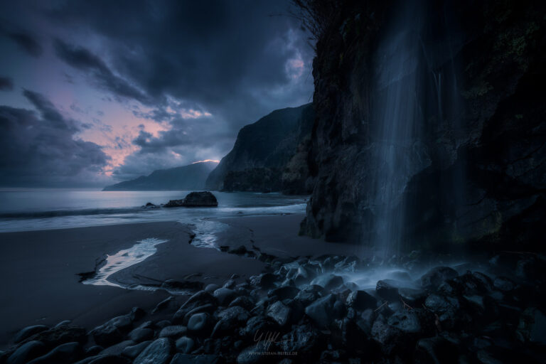Dark blue morning scene on the north coast of Madeira with a waterfall on a black sand beach and mystical dawns